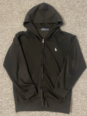 Svart zip hoodie från Polo Ralph Lauren - Snygg svart hoodie med dragkedja från Polo Ralph Lauren. Klassisk design med huva, fickor framtill och den ikoniska vita logon broderad på bröstet. Perfekt till chill dagar och enkel att matcha med allt. Mjuk och skön bomullskänsla.