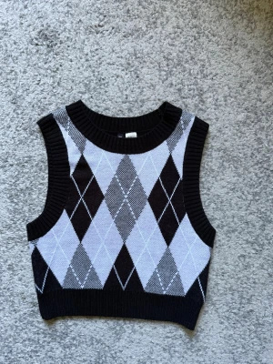 Svart och lila rutig stickad väst - Trendig stickad väst med svart och lila argyle-ruta. Croppad modell med bred ribbad kant vid hals och ärmhål. Perfekt att styla över en skjorta eller t-shirt för en cool look.