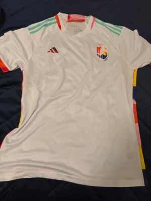 Belgien tröja  - Säljer en vit fotbollströja från Adidas i storlek L. Tröjan har färgglada detaljer längs sidorna och axlarna, samt tryckta loggor på bröstet. Slim fit-modell i lätt och bekvämt material, perfekt för träning eller match.