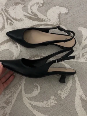 Svarta slingback pumps med låg klack - Stilrena svarta pumps med spetsig tå och slingback-rem. Skorna har en låg, smal klack och justerbart spänne vid ankeln. Tillverkade i ett slätt material som ger en elegant look. Perfekta för dig som gillar klassisk och trendig stil.