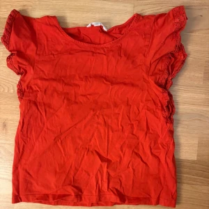 Röd topp med spetsdetaljer - Snygg röd topp med korta ärmar och spetsdetaljer längs ärmsluten. Toppen har rund halsringning och är gjord i mjuk bomull, perfekt för en trendig och färgstark look.