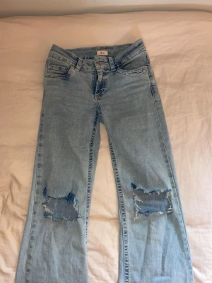 Ljusblå hål jeans med brodyr - Ljusblå bootcut jeans och slitningar på båda knäna. Baksidan har fickor med rosa och vit broderad zigzag-detalj. Jeansen har klassisk femficksmodell och är tillverkade i denim.