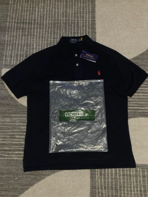 Marinblå piké från Polo Ralph Lauren - Klassisk marinblå pikétröja från Polo Ralph Lauren med korta ärmar, krage och två knappar framtill. Ikoniska röda loggan broderad på bröstet. Perfekt till jeans eller shorts för en clean och stilren look.