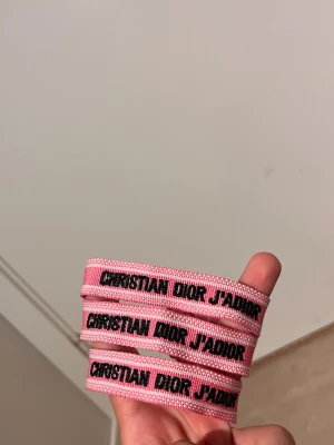 Christian Dior rosa textarmband - Säljer tre snygga rosa tygarmband från Christian Dior med svart text 'CHRISTIAN DIOR J'ADIOR'. Armbanden har en vävd struktur och justerbar knytning. Perfekt accessoar för dig som gillar exklusiva och trendiga detaljer.