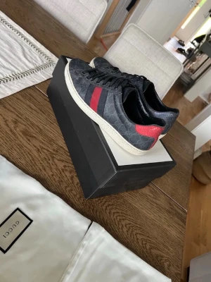 Gucci ace - Snygga svarta Gucci sneakers med klassiskt GG-mönster och röda samt blå ränder på sidan. Skorna har vita sulor och är låga i modellen. Materialet är en mix av canvas och läder, vilket ger en exklusiv känsla. Perfekta för dig som vill sticka ut med stil. Storlek 42 du kan skriva för mer bilder eller om du har frågor😁