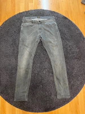 Gråa slim fit jeans från Lee - Säljer ett par gråa slim fit jeans från Lee med klassisk femficksdesign och knappgylf. Jeansen har en tvättad look och raka ben. Perfekta för dig som gillar en avslappnad men ändå stilren vibe.