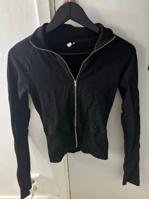 Svart zip hoodie - Säljer en svart kofta med dragkedja framtill. Modellen är figurnära med långa ärmar och ribbade muddar vid ärmslut och nederkant. Perfekt till chill eller streetwear-stil. Materialet känns mjukt och stretchigt.