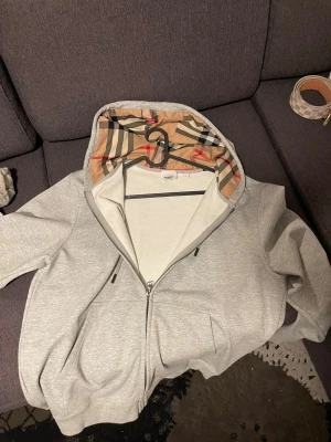 Grå zip hoodie med Burberry-mönster - Snygg grå zip hoodie från Burberry med klassiskt rutigt mönster i huvan. Hoodien har dragkedja framtill, två fickor och justerbar dragsko i huvan. Perfekt för en chill och stilren look. Materialet är mjukt och bekvämt, och loggan syns diskret på bröstet.