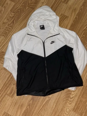Svartvit vindjacka från Nike - Snygg vindjacka från Nike i svart och vitt med dragkedja framtill och huva. Jackan har två sidofickor med dragkedja och klassisk Nike-logga på bröstet. Perfekt för blåsiga dagar och sportiga outfits.