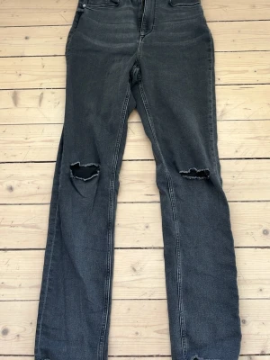 Svarta raka jeans med hål på knäna - Snygga svarta jeans med raka ben och slitna hål på båda knäna. Jeansen har hög midja, klassiska bakfickor och är tillverkade i stretchigt denimtyg. Perfekta för en avslappnad och trendig stil.