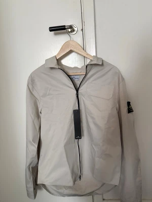 Beige overshirt från Stone Island - Snygg beige overshirt från Stone Island med dragkedja framtill, klassisk krage och en bröstficka. Ikonisk logopatch på ärmen. Tillverkad i ett lätt material som passar perfekt för lager-på-lager. Enkel och stilren design som funkar till det mesta.