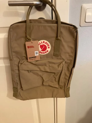 Beige Kånken ryggsäck från Fjällräven - Klassisk beige Kånken ryggsäck från Fjällräven med ikonisk röd logotyp framtill. Ryggsäcken har ett stort huvudfack, en framficka med dragkedja och justerbara axelband i slitstarkt material. Helt ny, säljer då jag fick den i pris från skolan utan kvitto och har tyvärr ingen användning för den.