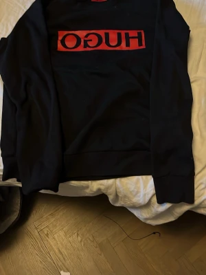 Svart HUGO hoodie med röd logga - Snygg svart hoodie från HUGO med stor röd logga på bröstet. Klassisk passform med långa ärmar och ribbade muddar. Perfekt för dig som gillar streetwear och vill ha en enkel men stilren look.