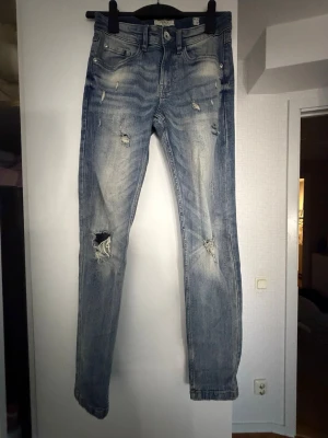 Ljusblå slitna jeans med hål - Säljer ett par ljusblå jeans med slitningar och hål på knäna. Jeansen har fem fickor, normal midja och smal passform. De är tillverkade i denim och har en cool, tvättad look med tydliga slitdetaljer framtill.