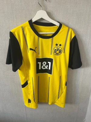 Borussia Dortmund matchtröja Puma - Gul och svart Borussia Dortmund matchtröja från Puma med klubbmärke, sponsorlogga och svarta detaljer. Klassisk rund halsringning och korta ärmar. DORTMUND tryckt på ryggen. Perfekt för fotbollsfans och träning.