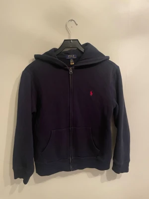 Mörkblå hoodie från Polo Ralph Lauren - Mörkblå hoodie med dragkedja från Polo Ralph Lauren. Klassisk design med huva och den ikoniska röda logotypen broderad på bröstet. Tillverkad i mjukt bomullsmaterial som är perfekt för chill dagar. Storleken är ganska liten och passar till 12-13 åringar. 