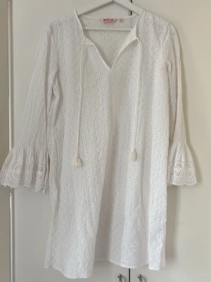 Vit strandtunika med broderie anglaise - Luftig vit strandtunika i broderie anglaise med v-ringning och knytband. Tunikan har långa ärmar med volangkant och är perfekt att dra över bikinin på stranden. Materialet är lätt och andas, vilket gör den skön under varma dagar.