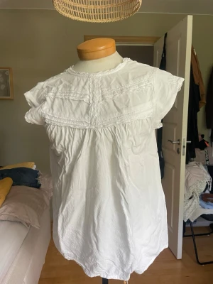 Vit blus med spetsdetaljer - Söt vit blus med korta ärmar och fina spetsdetaljer framtill. Blusen har en lös passform, liten volangkrage och knäppning bak i nacken. Perfekt till jeans eller kjol för en fräsch look. Passar jätte bra till sommaren men även till kallare väder med en kofta