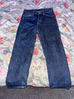 Mörkblå jeans med broderade fickor - Säljer ett par mörkblå jeans med raka ben och coola broderier på bakfickorna – ena fickan har en gul orm och den andra en vit drake. Jeansen har klassisk femficksdesign och kontrastsömmar. Perfekta för dig som vill sticka ut med detaljer! (Passar som w29)