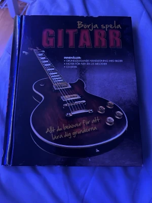Börja spela gitarr bok - En bok med titeln 'Börja spela gitarr' som innehåller grundläggande handledning med bilder, noter för fler än 25 melodier och CD-skiva. Omslaget är svart med en bild på en elgitarr och text i rött och gult. Perfekt för dig som vill lära dig spela gitarr från grunden.