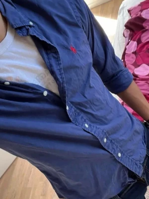 Polo Ralph Lauren skjorta  - Snygg blå skjorta med långa ärmar och klassisk krage. Skjortan har vita knappar framtill och en liten röd broderad logga på bröstet. Perfekt till jeans eller chinos för en clean look.