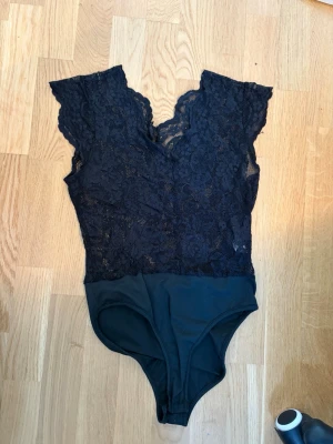 Svart spetsbody med v-ringning - Snygg svart body i spets med v-ringning och korta ärmar. Överdelen är helt i genombruten spets med blommönster och nederdelen i slät stretchig tyg. Perfekt för att skapa en trendig look med lite extra edge.