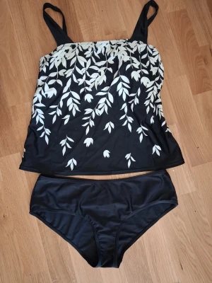 Svartvit tankini med bladmönster - Snygg svart tankini med vita blad och justerbara axelband. Klassisk modell med rundad halsringning och hel baksida. Mönstret ger en fräsch och stilren look, perfekt för stranden eller poolen. Troligtvis i stretchigt material som polyester eller elastan.