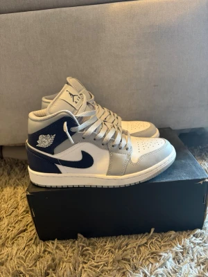 Nike Air Jordan 1 Mid grå/blå - Snygga Nike Air Jordan 1 Mid sneakers i grått, vitt och marinblått. Skorna har klassisk Jordan-logga på sidan och tungan, perforerad tåbox och vita snören. Tillverkade i läder och textil för en clean och sportig look.