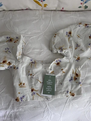 Vit blommig topp med puffärmar - Söt vit topp med blommigt mönster i lila, gult och brunt. Toppen har  puffärmar och en fyrkantig urringning. Köpt på hm och aldrig använd, lapparna kvar :)