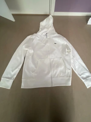 Vit Lacoste zipup helt ny size M perfekt till sommaren och svarta byxor eller jeans  - Snygg vit hoodie från Lacoste med dragkedja framtill och klassisk krokodil-logga på bröstet. Hoodien har huva med dragsko och två stora fickor framtill. Perfekt för en clean och stilren look. Materialet ser ut att vara mjukt och bekvämt.