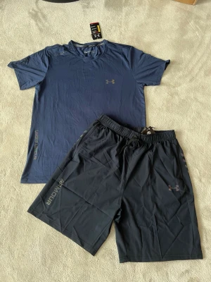 Under Armour träningsset t-shirt & shorts - Snyggt träningsset från Under Armour med en mörkblå t-shirt och svarta shorts. T-shirten har rund hals och diskret logga på bröstet, medan shortsen har elastisk midja och logga på benet. Perfekt för gymmet eller löprundan.