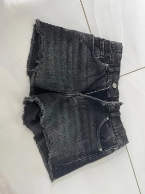 Svarta jeansshorts med rå kant - Säljer ett par svarta jeansshorts med fransig, rå kant och klassisk femficksdesign. Shortsen har dragkedja och knapp framtill samt bälteshällor. Perfekta för sommaren och ger en avslappnad vibe.