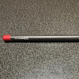 MAC Läppenna Brick röd - MAC Lip Pencil i nyansen Brick, en klassisk röd läppenna med matt finish. Pennan har svart kropp med vit logga och färgad ände som visar nyansen. Perfekt för att definiera och forma läpparna med precision.