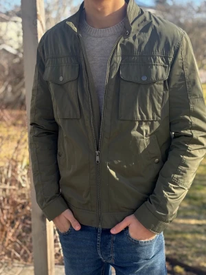 Olivgrön Vindjacka/Fieldjacka - Snygg olivgrön vindjacka/fieldjacka med dragkedja och tryckknappar. Jackan har två stora bröstfickor med lock och två sidofickor. Ståkrage med knapp och raka ärmar. Perfekt för dig som vill ha en stilren och funktionell jacka till vardags. Materialet är lätt och vindtätt.  Jackan är Storlek M men är lite mindre i storleken.