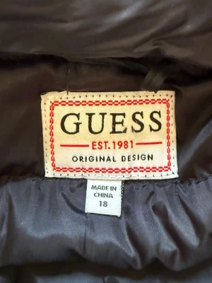 Svart dunväst från Guess - Snygg svart dunväst från Guess med quiltad design och hög krage. Västen har dragkedja framtill, två fickor med dragkedja och en liten Guess-logga på bröstet. Perfekt för lager-på-lager och enkel att matcha till olika outfits. Lite sönder inuti en ficka