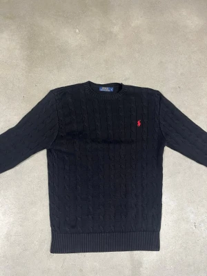 Svart stickad Polo Ralph Lauren - Kabelstickad svart tröja från Polo Ralph Lauren med rund halsringning och röd broderad logga på bröstet. Tröjan har långa ärmar och ribbade muddar vid ärmslut och nederkant. Perfekt för en stilren och klassisk look. Jag är 193 och tröjan passar utmärkt, så räkna med att tröjan passar dig som även har M/L.