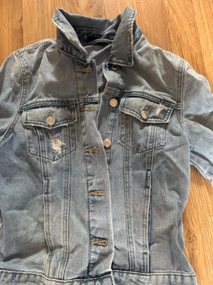 Ljusblå klassisk jeansjacka - Snygg ljusblå jeansjacka med slitna detaljer och silverfärgade knappar. Två bröstfickor med lock och knapp, samt markerad krage. Jackan har en croppad passform. Perfekt för en cool och avslappnad stil.