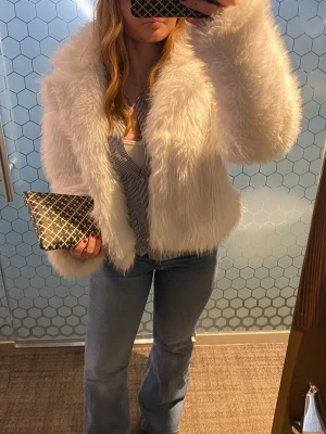 Vit fluffig pälsjacka - Säljer en supermjuk och fluffig vit pälsjacka med bred krage och rak passform. Jackan har en lyxig look och är perfekt för att hålla värmen under kalla dagar. Passar dig som vill sticka ut och addera lite glam till din outfit.