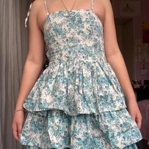 Blommig blå-vit kortklänning - Söt kortklänning med blått och vitt blommönster, smala axelband med knyt och-väldigt puffiga-volangdetaljer i kjolen. Klänningen har en feminin siluett och är perfekt för varma dagar. Tillverkad i mjukt bomullstyg som känns skönt mot huden. Axelbanden är justerbara 👍