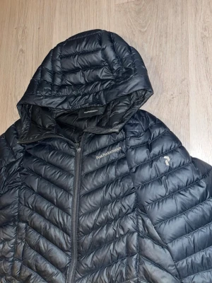 Svart dunjacka från Peak Performance - Snygg svart dunjacka från Peak Performance med huva och dragkedja framtill. Jackan har ett quiltat mönster och logga på bröstet samt ärmen. Perfekt för kalla vinterdagar och riktigt lätt att bära.