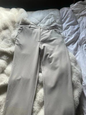 Beige kostymbyxa med raka ben - Snygga beige kostymbyxor med raka ben och pressveck. Byxorna har hällor för skärp, sidofickor och stängs med knapp och dragkedja. Perfekta för en clean och stilren look. Materialet ser ut att vara en mjuk och lätt polyesterblandning.