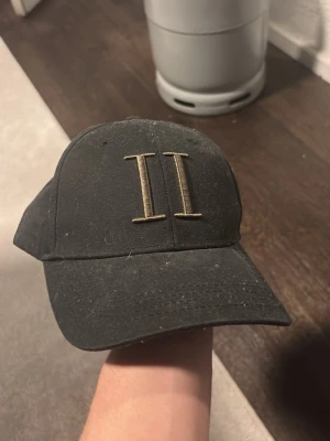 Svart keps från Les Deux - Svart keps från Les Deux med broderad logga i form av två romerska tvåor (II) framtill. Justerbar snapback baktill med liten Les Deux-tag. Klassisk böjd skärm och stilren design, perfekt för en clean streetwear-look.