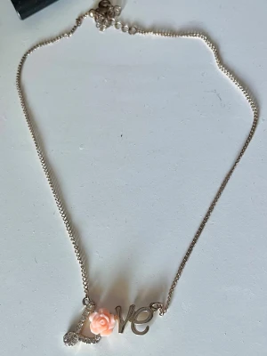 Guldfärgat halsband med LOVE och ros - Sött halsband i guldfärg med texten 'LOVE' som hänge. Bokstaven O är utformad som en ljusrosa ros och ” L” är klätt i ”diamanter”. Kedjan är tunn och smidig, perfekt för att ge en romantisk touch till din outfit.