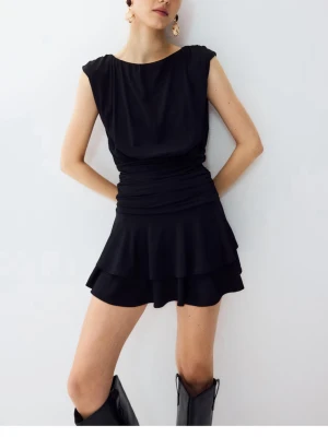 Svart kort klänning  - Säljer en svart kort klänning med rund halsringning och breda axelband. Klänningen har en figurnära midja med rynkdetaljer och en volangig kjol i två lager. Materialet ser ut att vara mjukt och följsamt, perfekt för en trendig look. HELT NY oanvänd säljer pga jag ej kunde returnera.