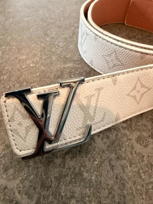 Vitt Louis Vuitton skärp i skinn - Snyggt vitt skärp från Louis Vuitton med klassiskt monogrammönster och stor LV-logga i silver på spännet. Tillverkat i skinn med exklusiv känsla och stilren design. Perfekt accessoar för att lyfta din outfit.