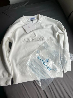 Vit Moncler sweatshirt topp - Kan gå ner i pris! Vit sweatshirt från Moncler med broderad logga och emblem på ärmen. Klassisk rund halsringning och ribbade muddar. Tillverkad i mjuk bomull för en clean och stilren look.