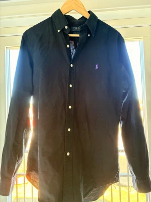 Svart skjorta från Polo Ralph Lauren - Klassisk svart skjorta från Polo Ralph Lauren i slim fit-modell. Skjortan har långa ärmar, vita knappar och den ikoniska lila logotypen broderad på bröstet. Tillverkad i mjuk bomull som känns skön mot huden. Perfekt för en stilren look.                             Säljer denna snygga skjorta eftersom den va lite stor på mig. Använt 1 gång. Skicket är grymt som nyskick. Tveka inte på att höra av er vid frågor🤩😁