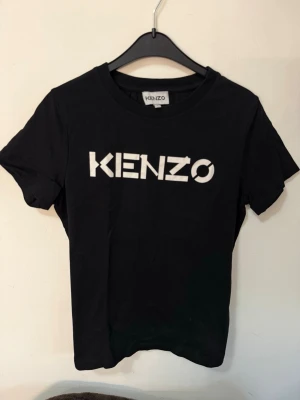 Svart Kenzo t-shirt med logga - Säljer en svart t-shirt från Kenzo med stor vit logga framtill. Klassisk rund halsringning och korta ärmar. Tillverkad i mjuk bomull för skön passform. Perfekt för dig som gillar stilrena och trendiga plagg med tydlig branding. Köpt för 999kr 