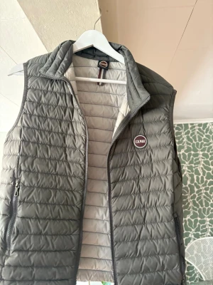 Grå dunväst från Colmar - Snygg grå dunväst från Colmar med quiltad design och hög krage. Västen har dragkedja framtill, två sidofickor med dragkedja och Colmar-logga på bröstet. Perfekt för lager-på-lager och ger en sportig vibe till din outfit.