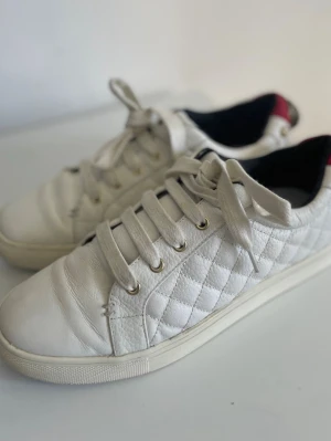 Vita läder sneakers New Balance 38 - Snygga vita sneakers från New Balance  med quiltad läderdesign och guldfärgade nitar. Skorna har röda detaljer på hälen med en unik metallapplikation och pärlor. Klassisk låg modell med vita snören och svart innerfoder. 